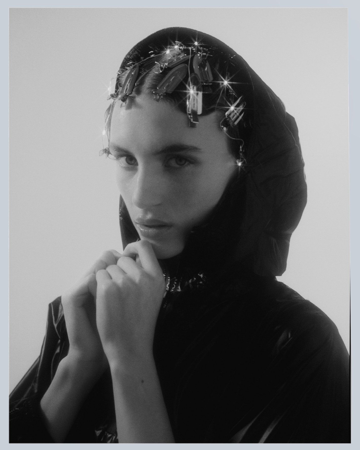 Schwarzweißfoto einer Person mit dunkler Kapuze, die die Hände am Kinn hält und eine glitzernde Kopfbedeckung trägt.