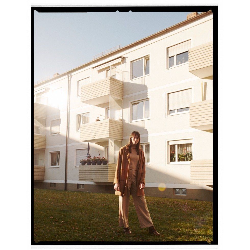 Frau im braunen Outfit steht vor einem Wohnhaus. Sonniger Tag, grünes Gras.