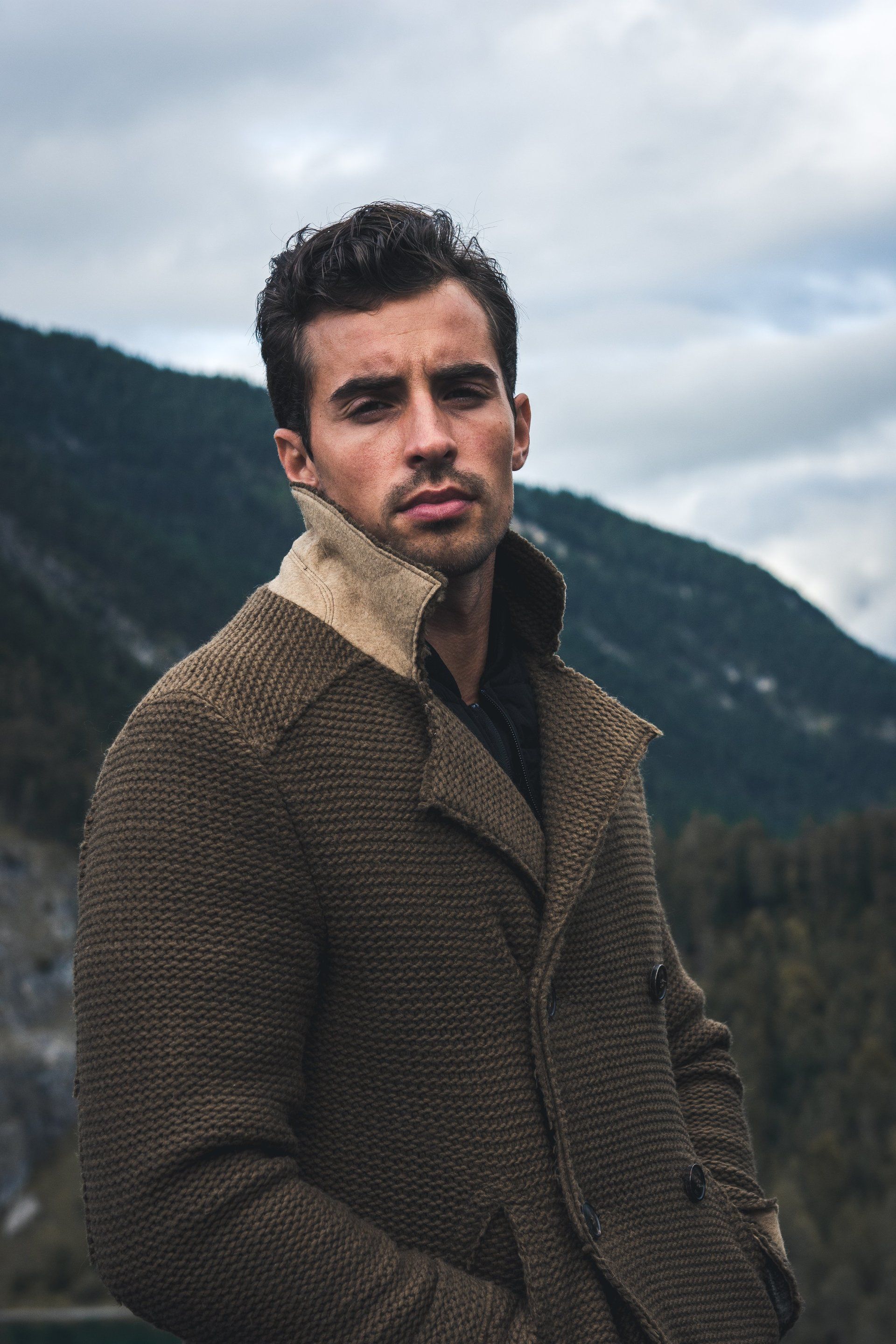 Mann in strukturiertem braunen Mantel steht an einem See mit Bergen im Hintergrund.