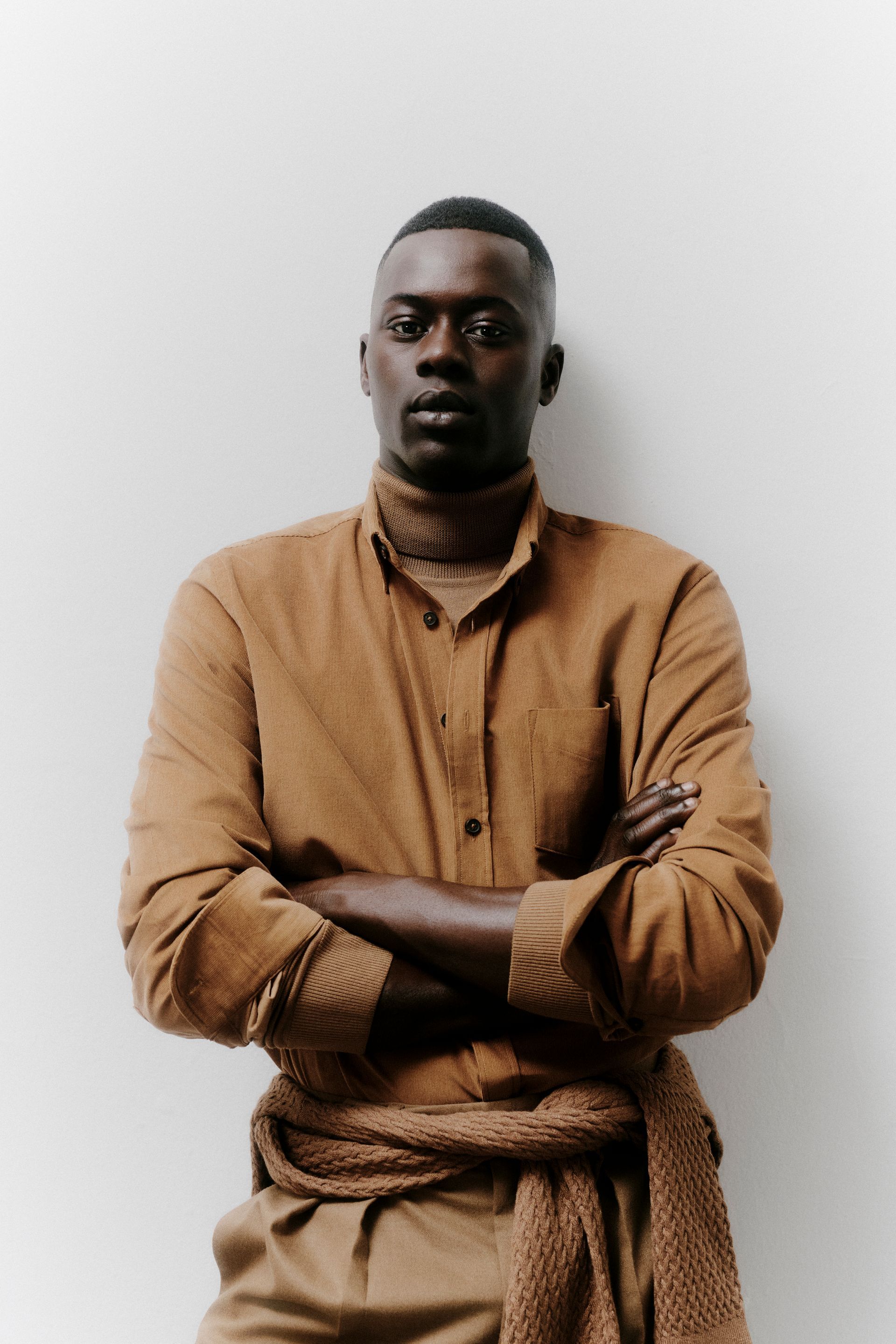 Mann mit verschränkten Armen, braunem Hemd und Rollkragenpullover, steht vor weißer Wand.