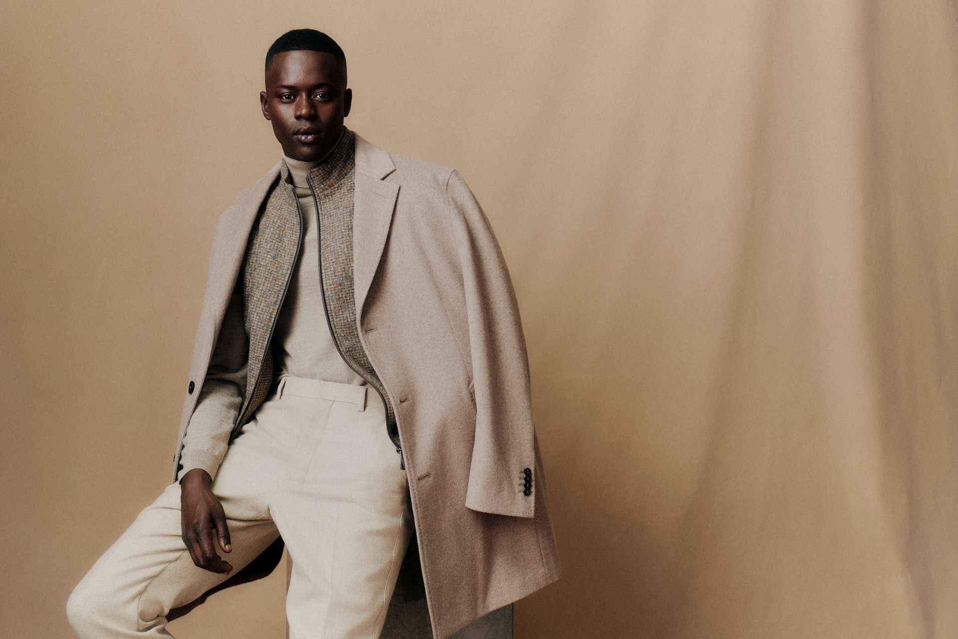 Mann in beigem Outfit und Mantel posiert auf schlichtem Hintergrund.