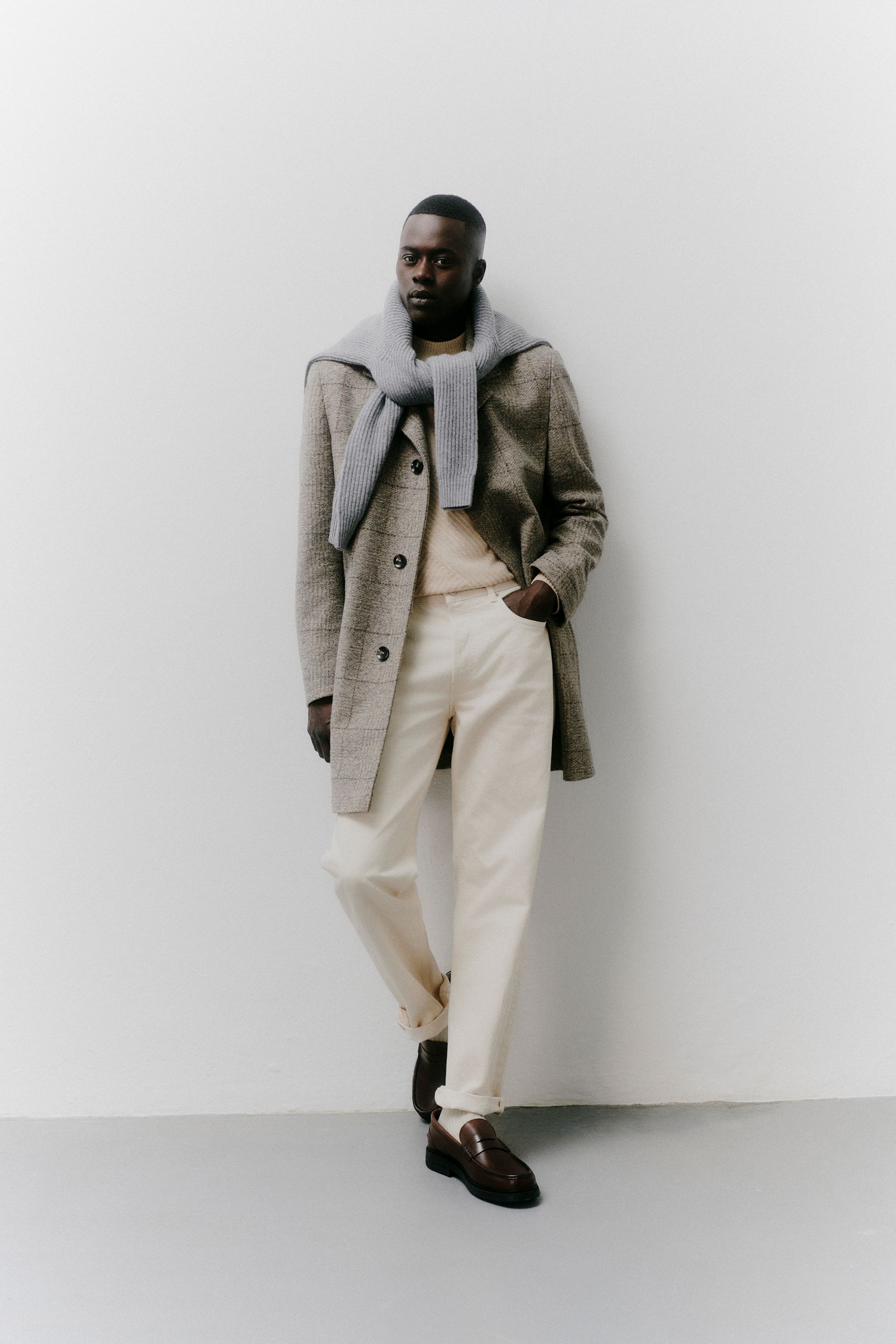 Mann mit gemustertem Mantel, cremefarbenen Hosen und Pullover lehnt an einer weißen Wand.