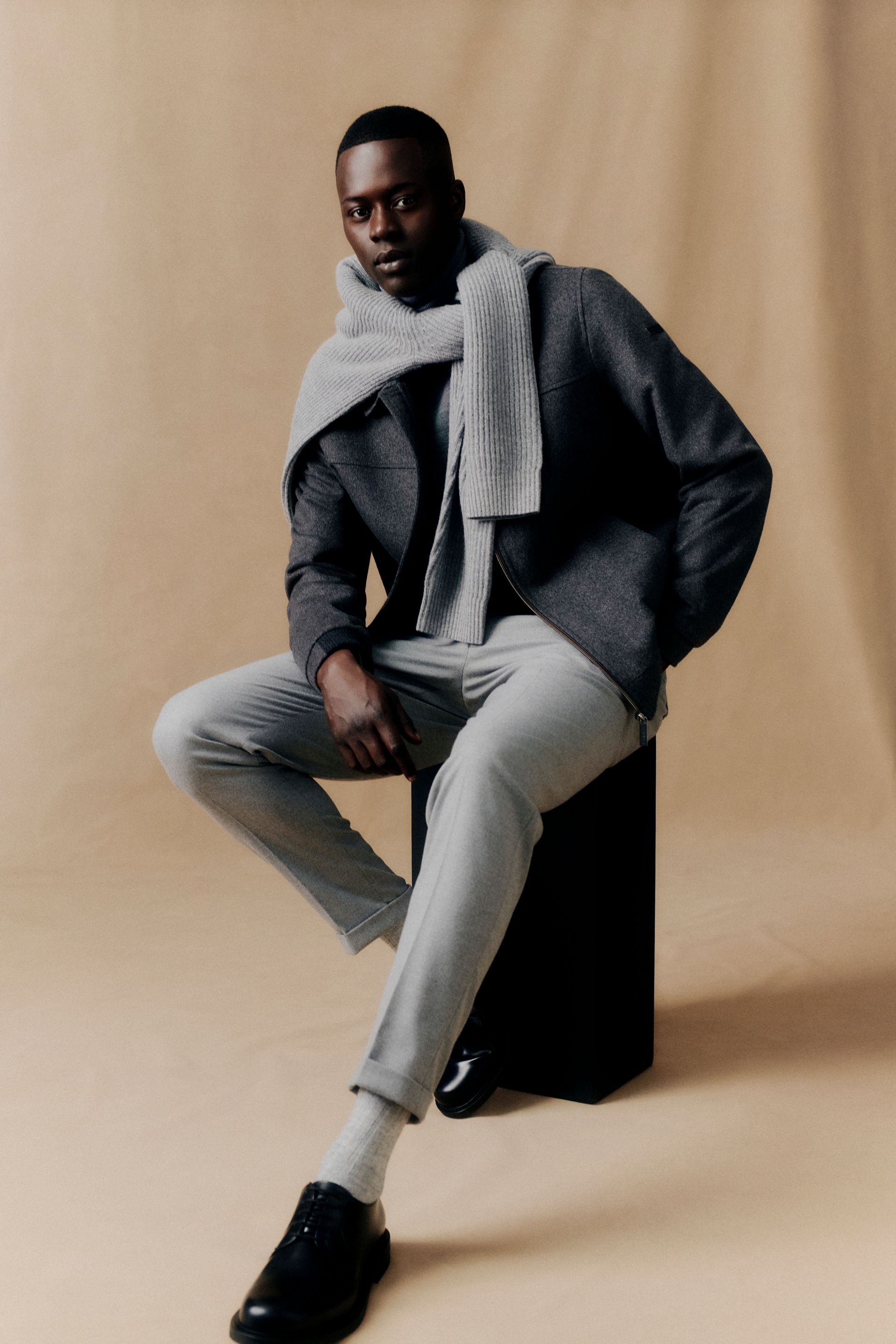 Mann in grauem Outfit, Schal, sitzend, schwarze Schuhe, neutraler Hintergrund.