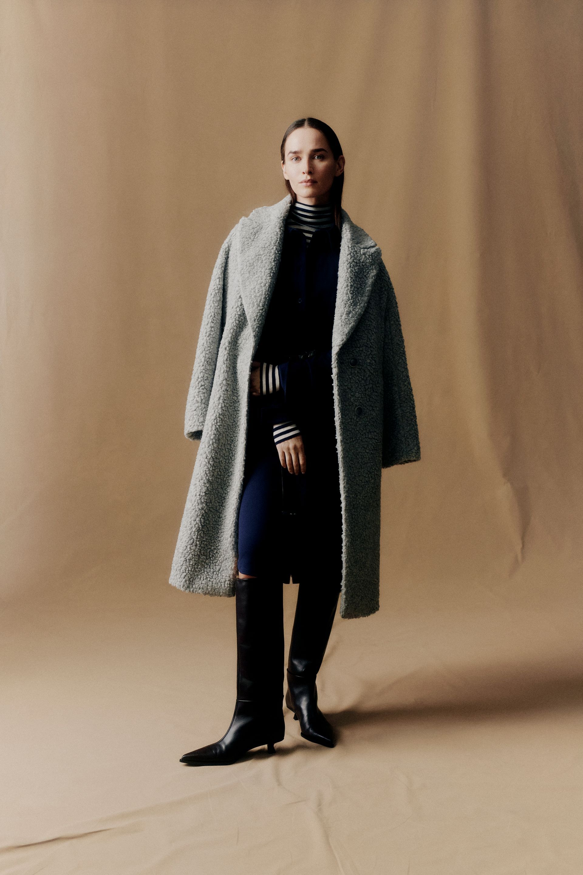 Frau in einem flauschigen grauen Mantel, dunklen Hosen und Stiefeln, posiert vor einem hellbraunen Hintergrund.