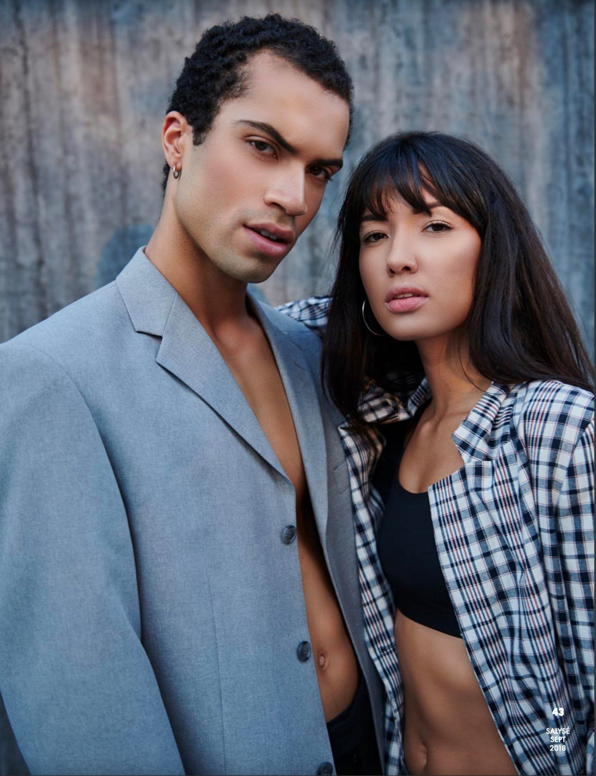 Mann im grauen Blazer und Frau im offenen karierten Hemd posieren vor einer Holzwand.