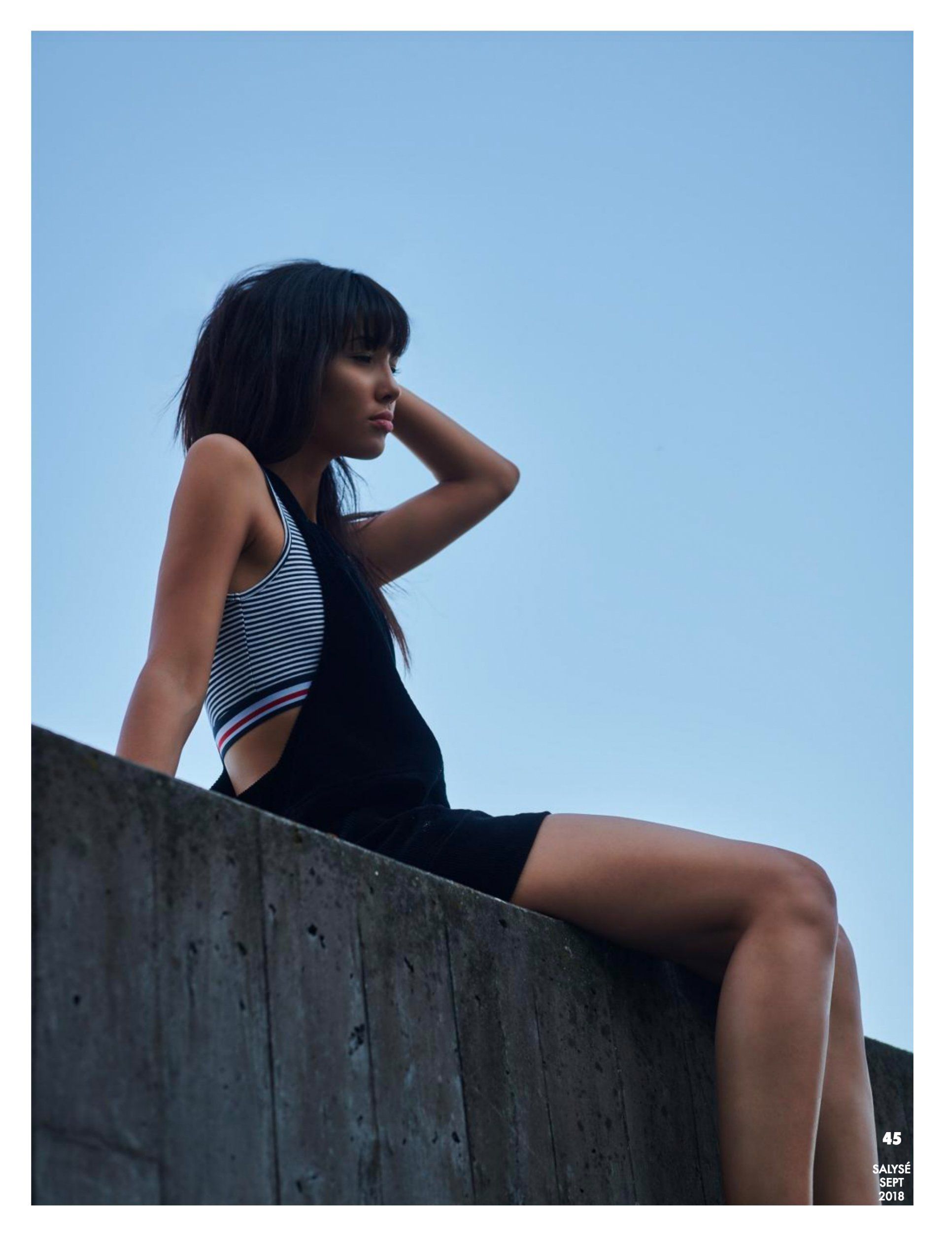 Frau in schwarzen Shorts und gestreiftem Oberteil sitzt auf Betonmauer und schaut weg. Hintergrund blauer Himmel.