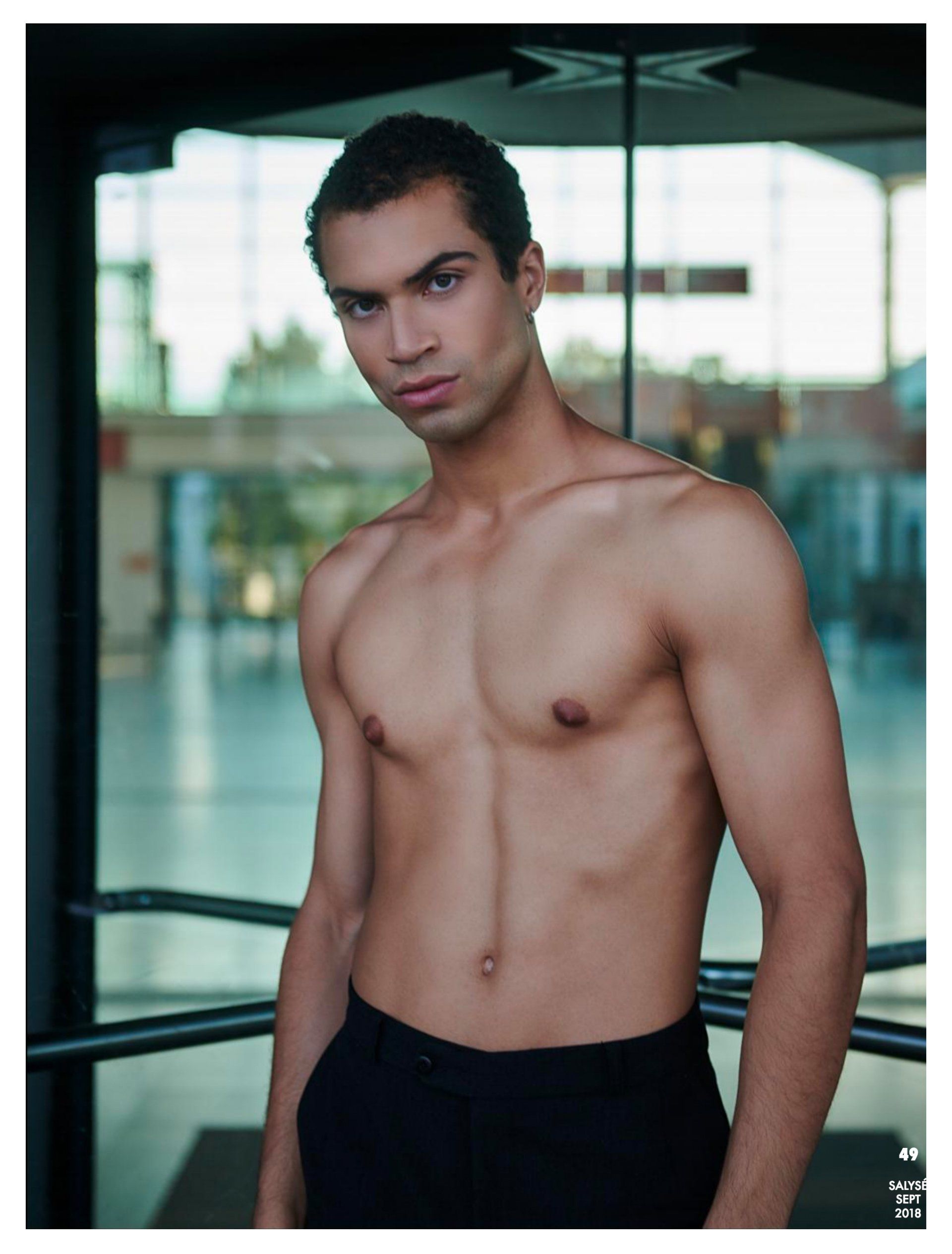 Ein Mann ohne Hemd schaut in die Kamera. Er trägt schwarze Hosen und sitzt in einem Gebäude mit Glastüren.