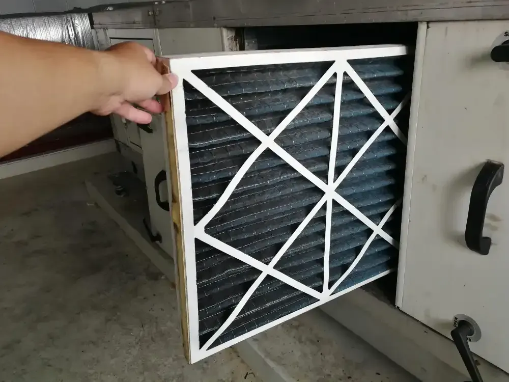 A hand pulling a used air filter from an HVAC unit. Filter is dark with a white frame.