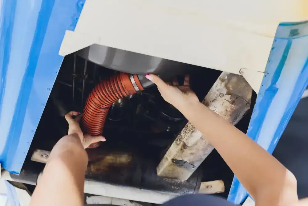Hands connecting an orange flexible hose to a metal component, likely in a vehicle, under a white structure.