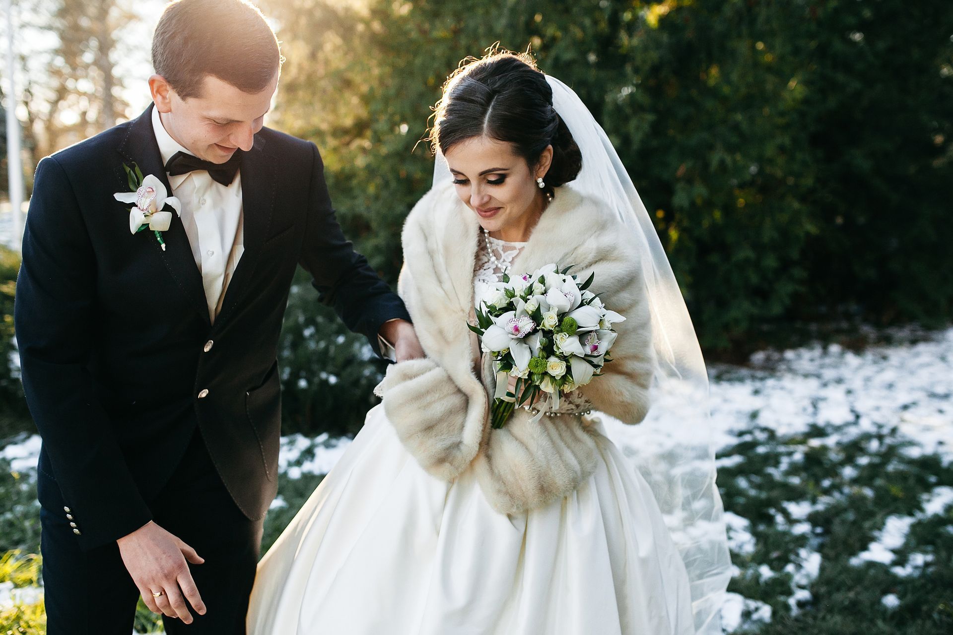 Groom helping bride wearing fur shawl exit ceremony, holding bouquet. Snowy outdoor setting, sunlight.