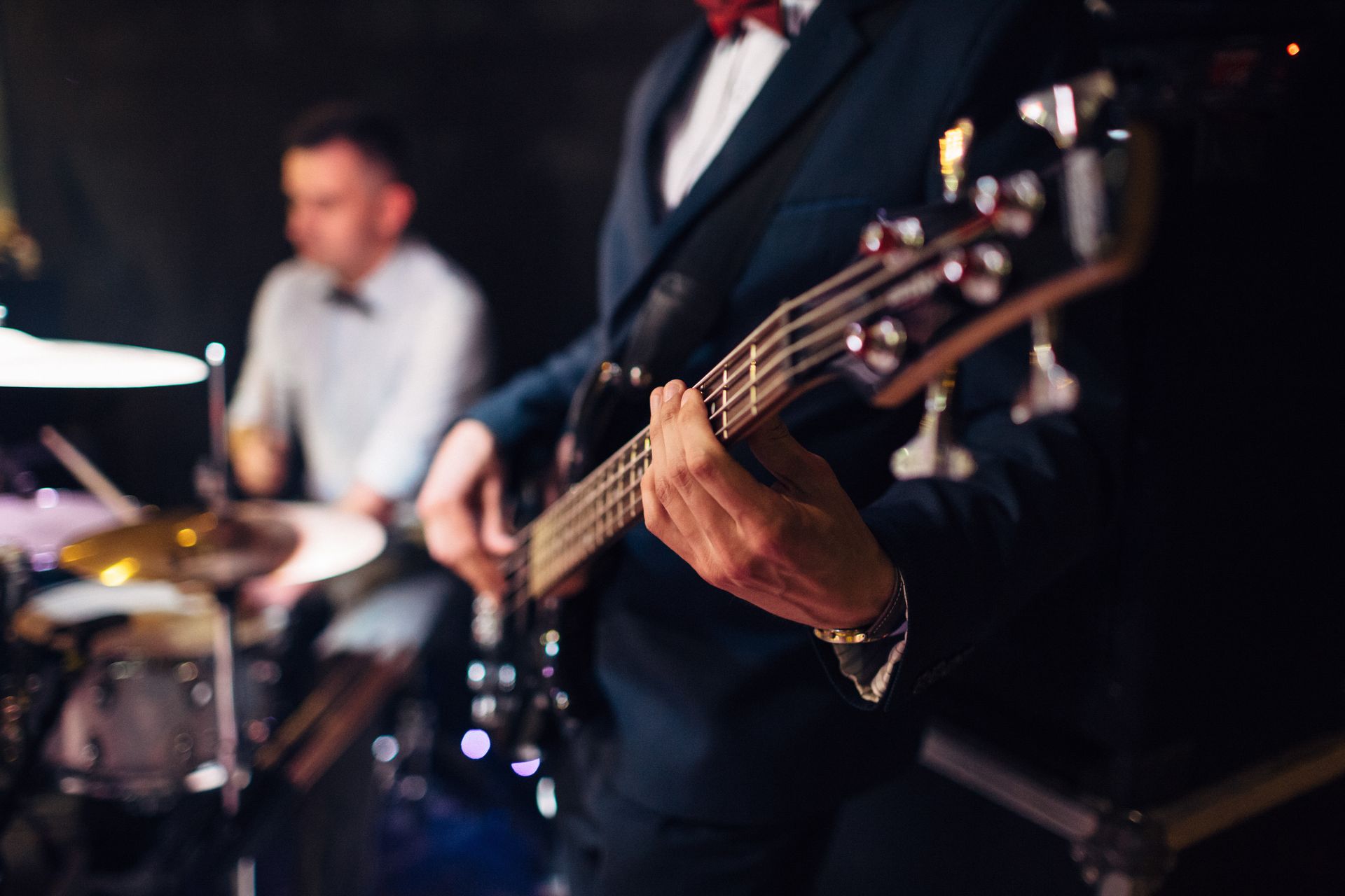 Man playing a bass guitar onstage, drummer in the background, dark setting.
