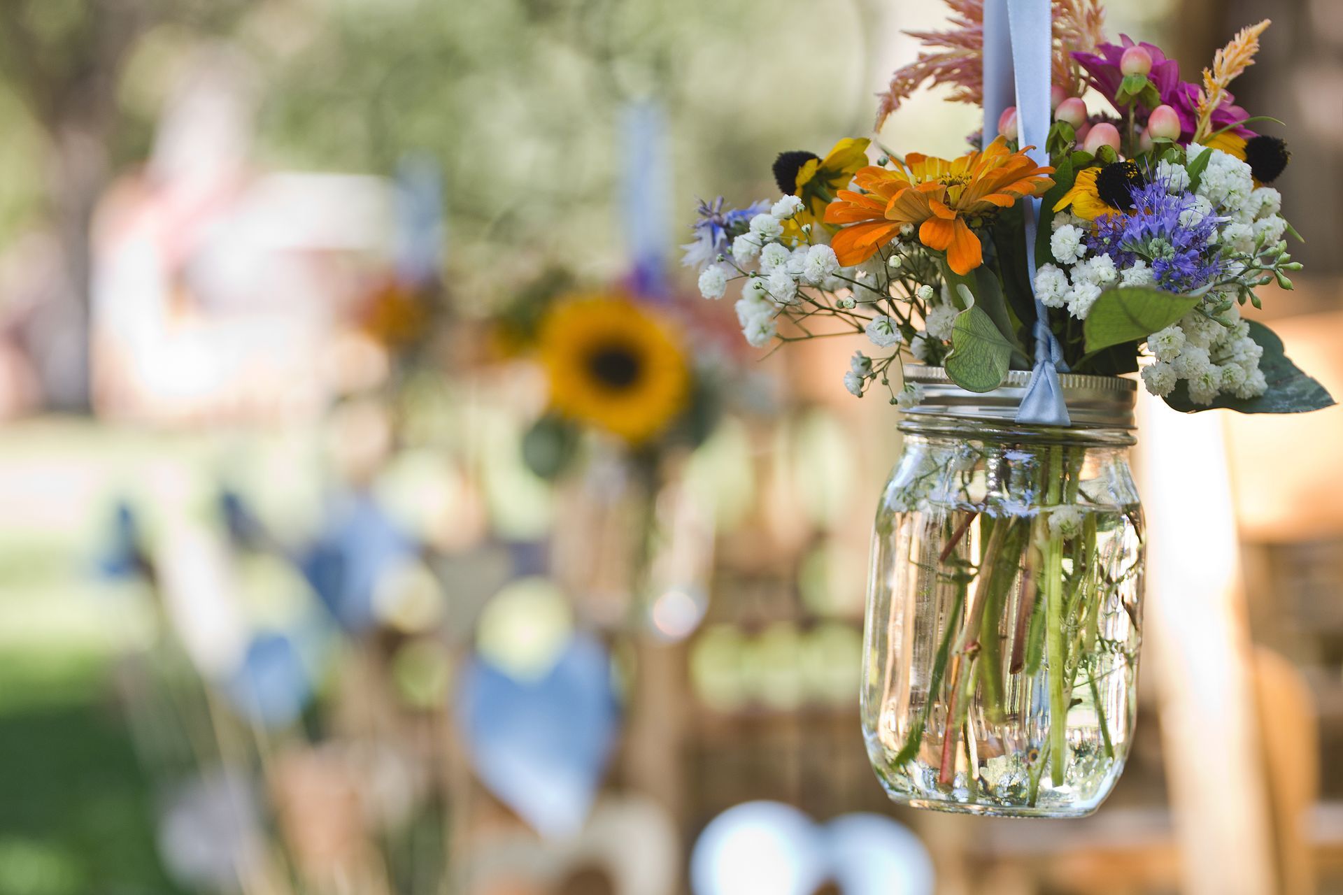 Mason jar with colorful flowers, hanging outdoors with blurred background of a sunny setting.