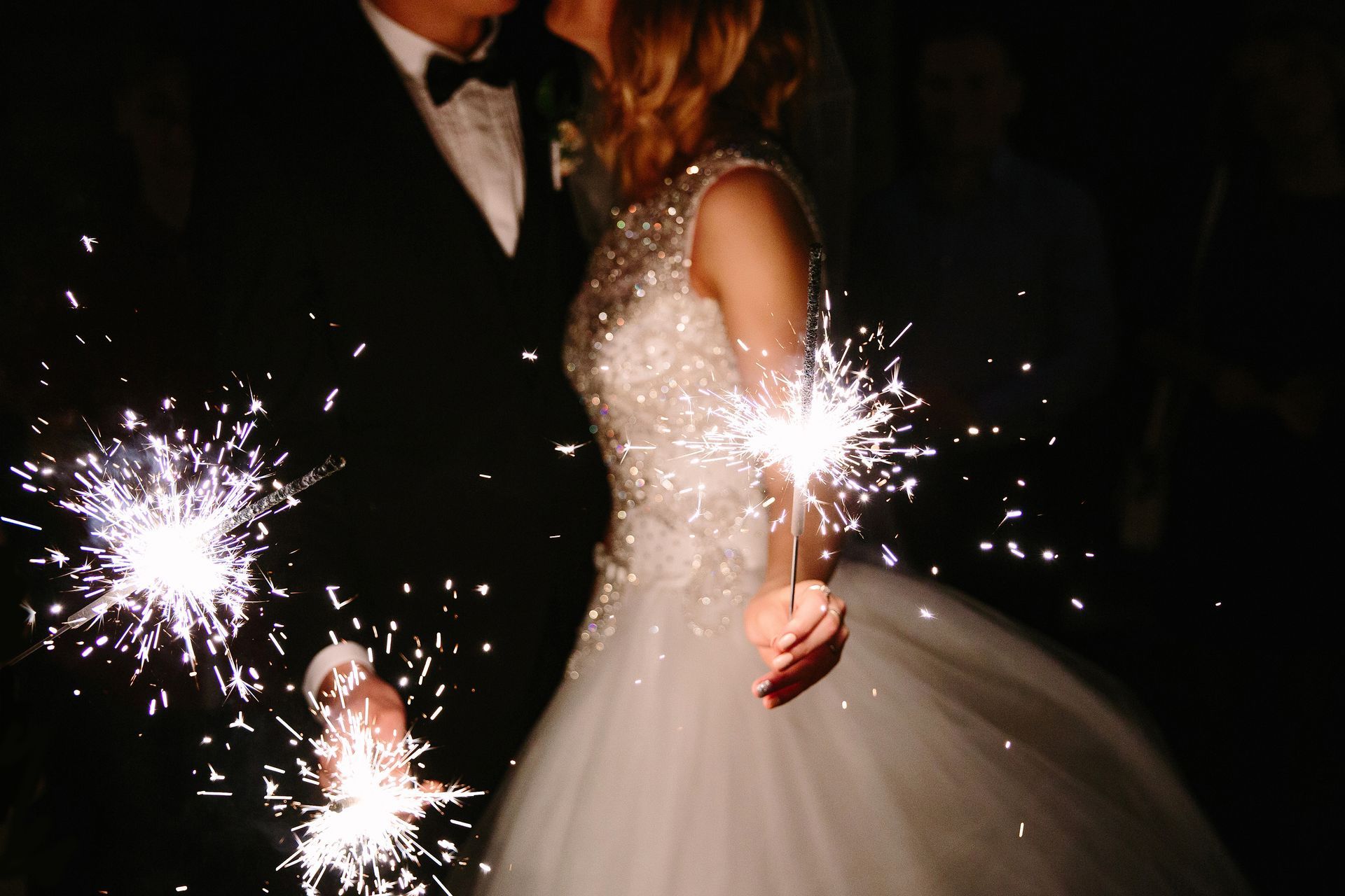 Couple kissing at a wedding; bride in a white gown and groom in a tuxedo with sparklers.