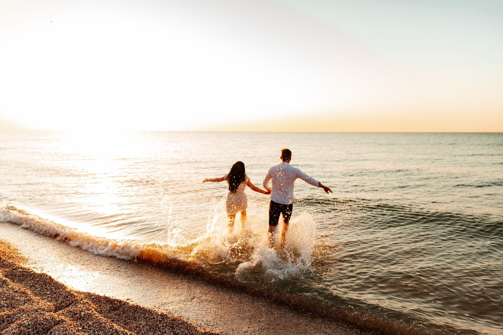 Couple holding hands and running into the ocean at sunset, water splashing around their feet.