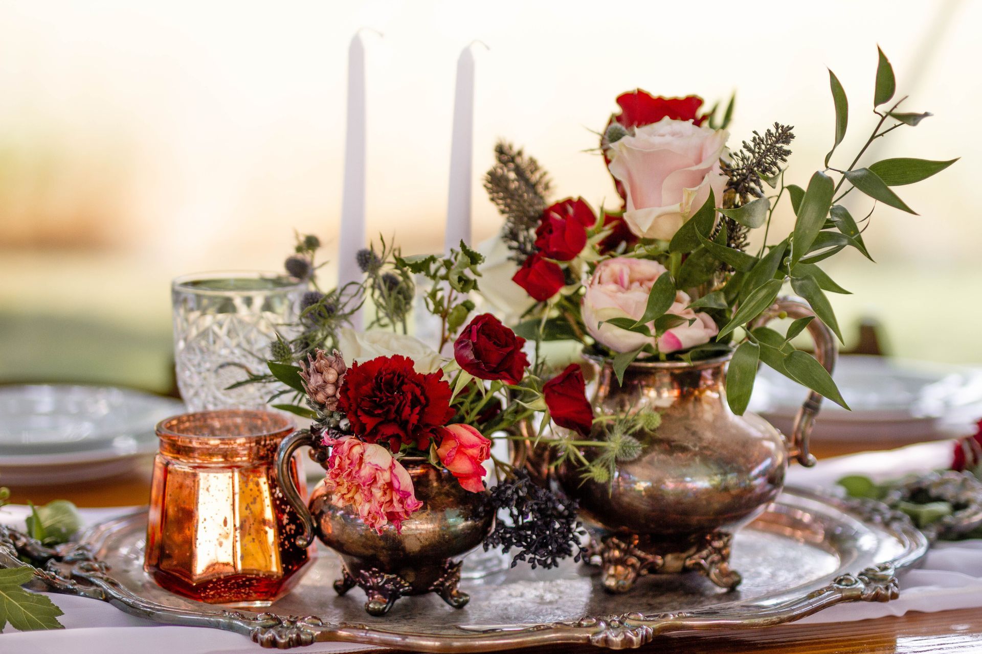 Floral centerpiece on silver tray with candles and a glass.