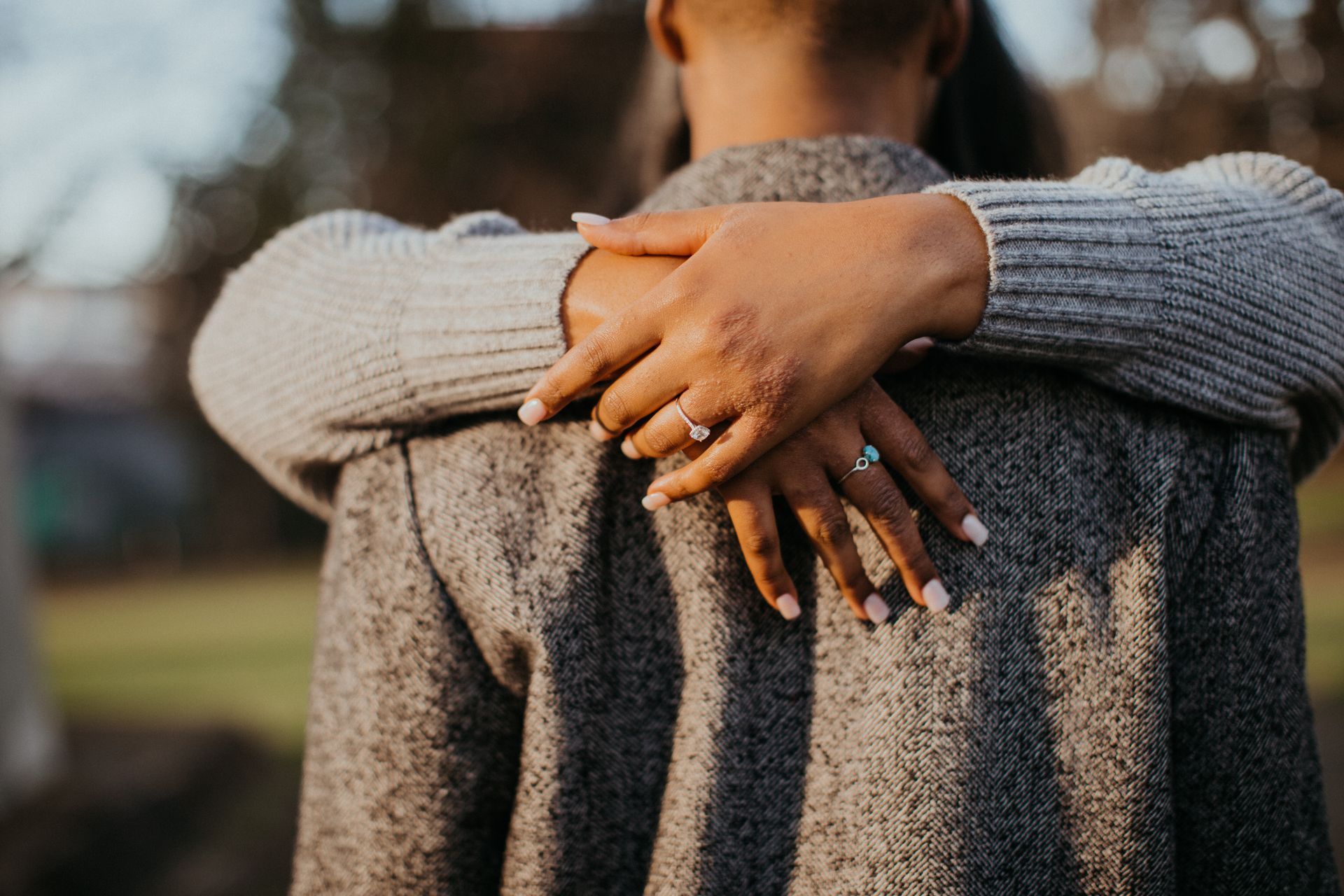 Hands with rings hug a person from behind. Gray sweater, blurred outdoor setting.