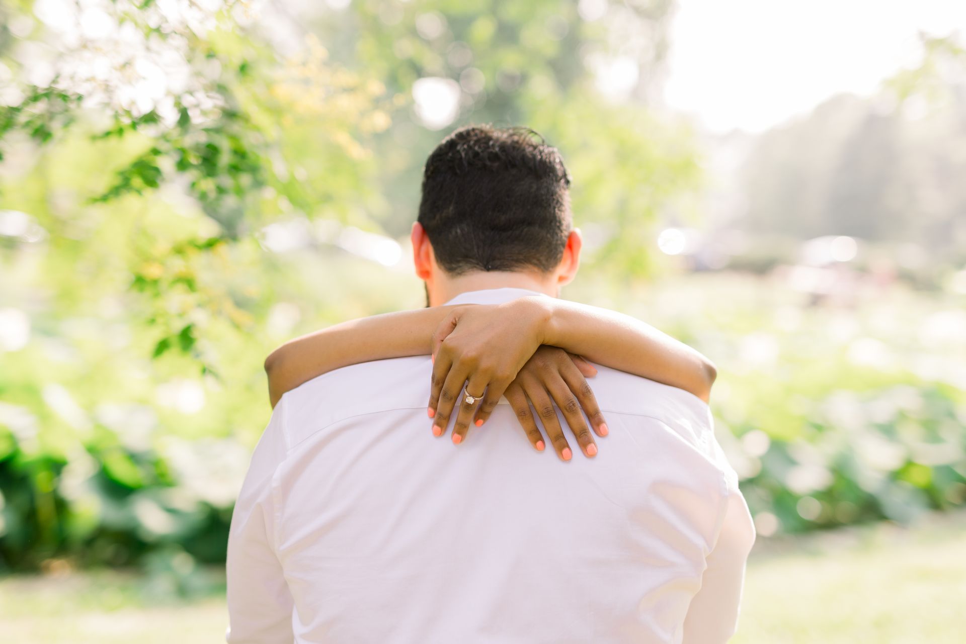 A man and a woman are hugging each other in a park. The woman is wearing a ring on her finger.