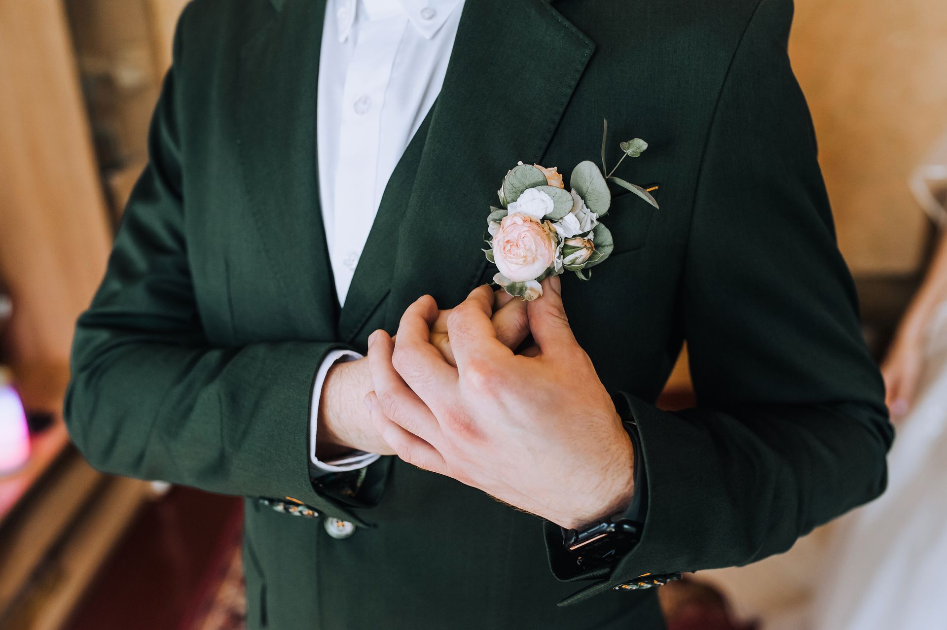 A groom in a dark green suit adjusting a boutonniere on his lapel. 