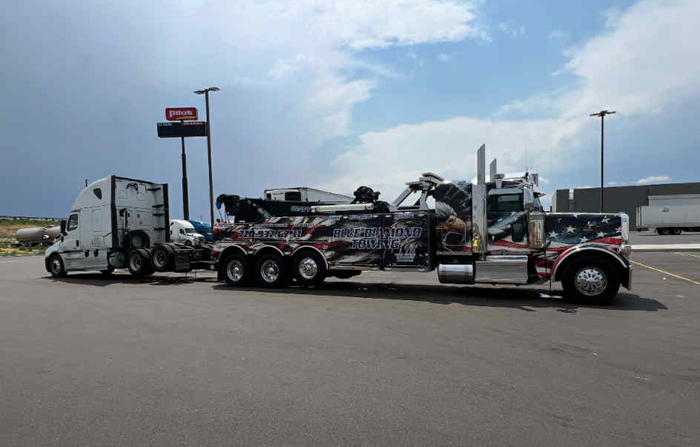A tow truck towing a semi-truck in a parking lot, under a cloudy sky.
