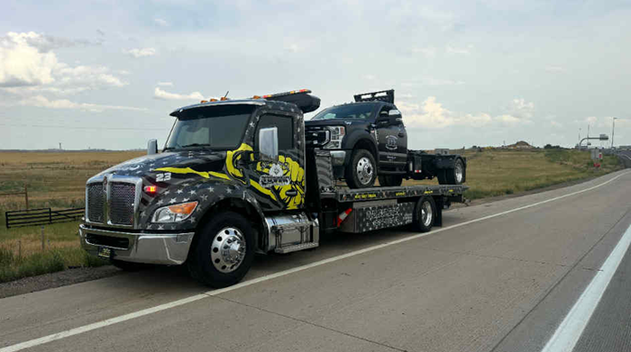 Tow truck on a highway, towing a black truck. The tow truck has a yellow and black design.