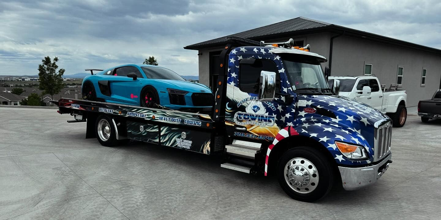 Blue sports car loaded on a tow truck, outside a building. The truck has a patriotic design.