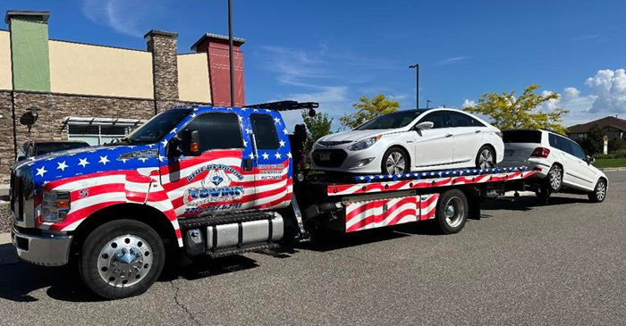 Tow truck with American flag design hauling two white cars on a sunny day.