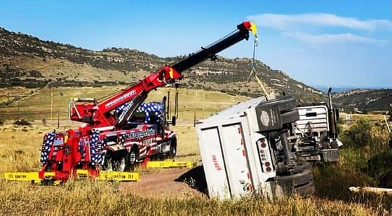 A large tow truck uprighting a white semi-truck overturned on a grassy hillside.