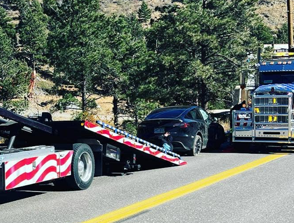 A black Tesla being loaded onto a tow truck on a road in a mountainous area, with trees in the background.