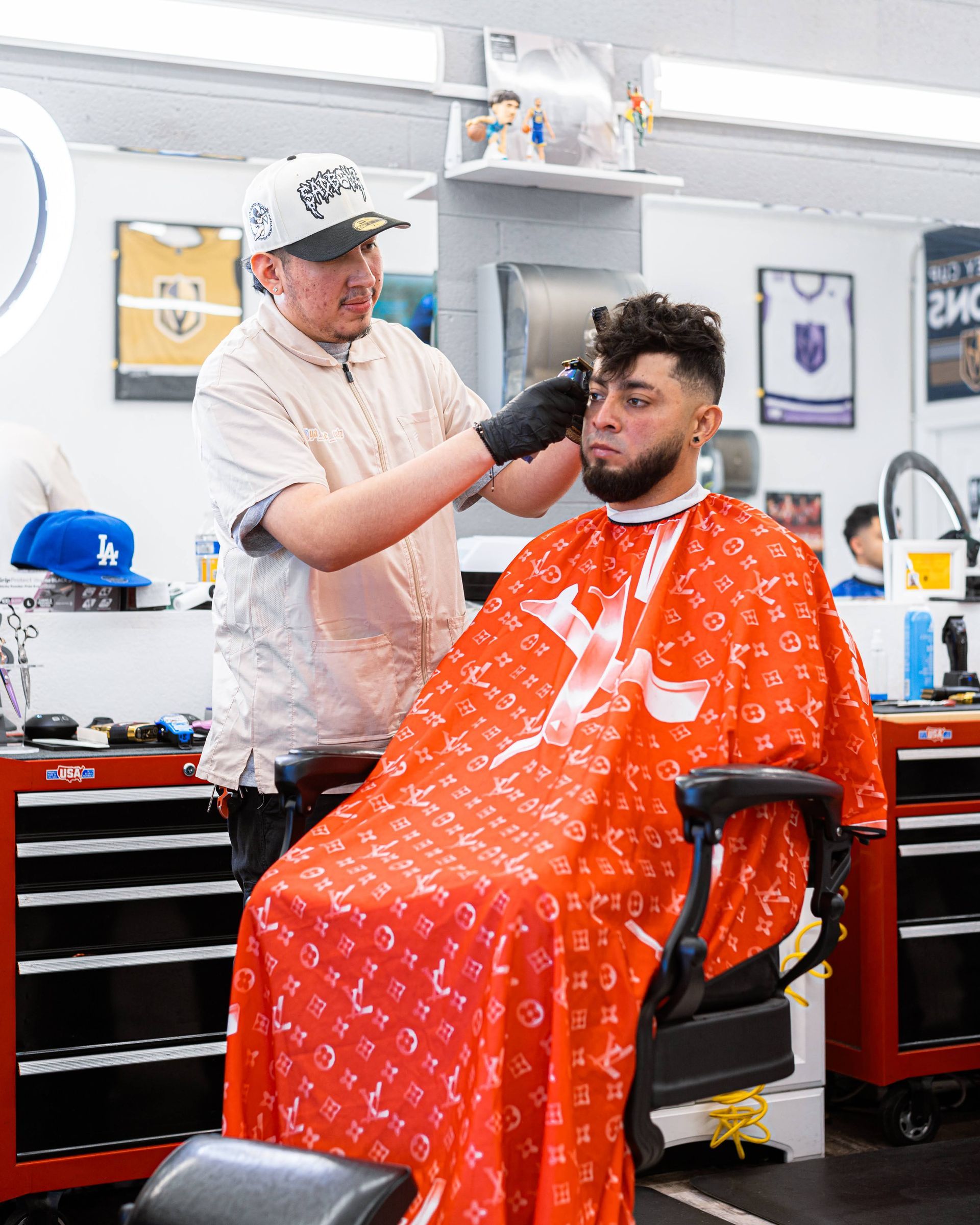 Barber trimming a client's beard in a barbershop, with the client seated in a red cape.