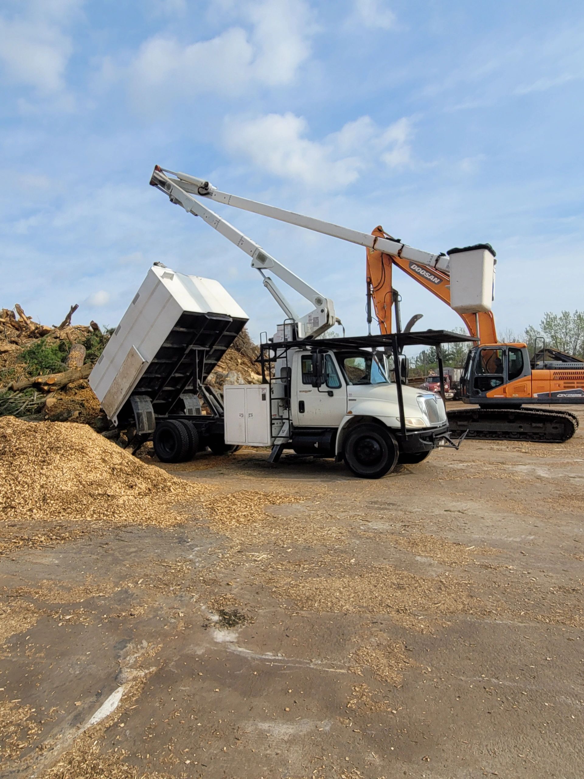 White truck with a lifted bed and boom in a lumberyard with an excavator.
