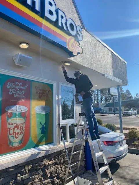 Man on a ladder painting a sign above a Dutch Bros coffee shop, sunny day.