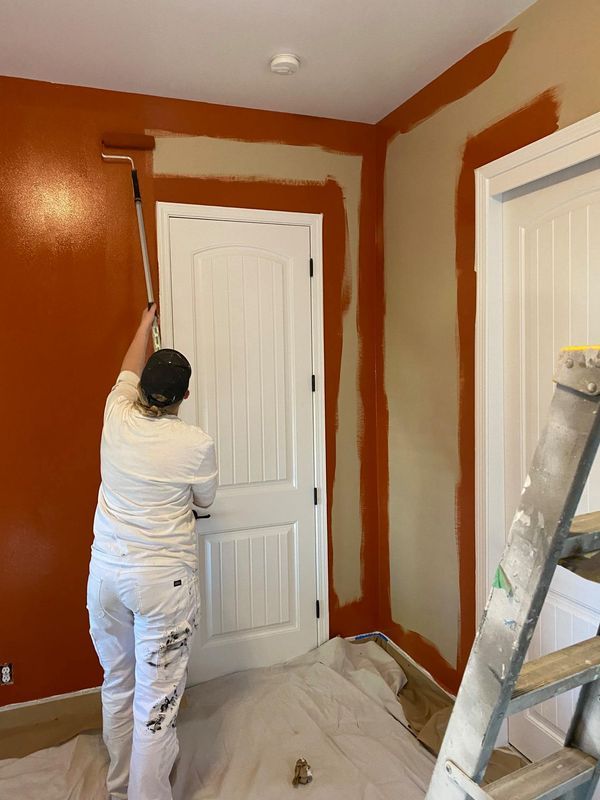 Person painting orange accent wall and trim in a room with white doors and beige walls, using a roller.