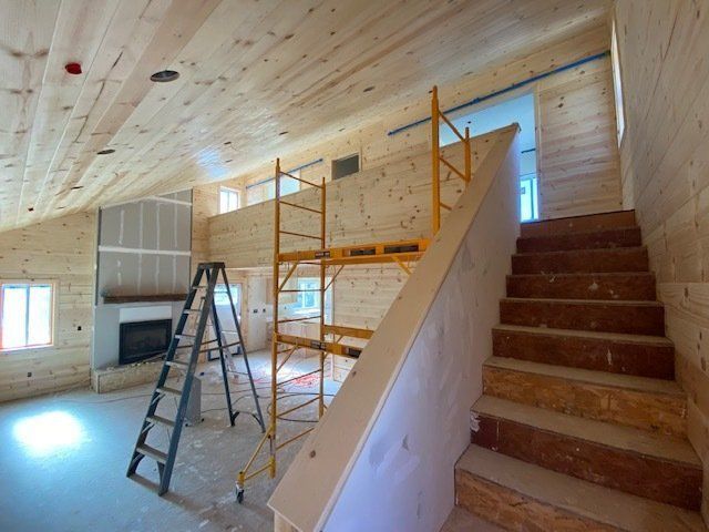 Interior view of a building under construction, featuring wooden walls, a staircase, scaffolding, and a fireplace.