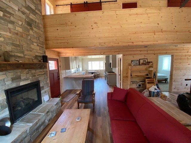 Cozy cabin interior with stone fireplace, wooden walls and ceiling, red couch, and open kitchen.