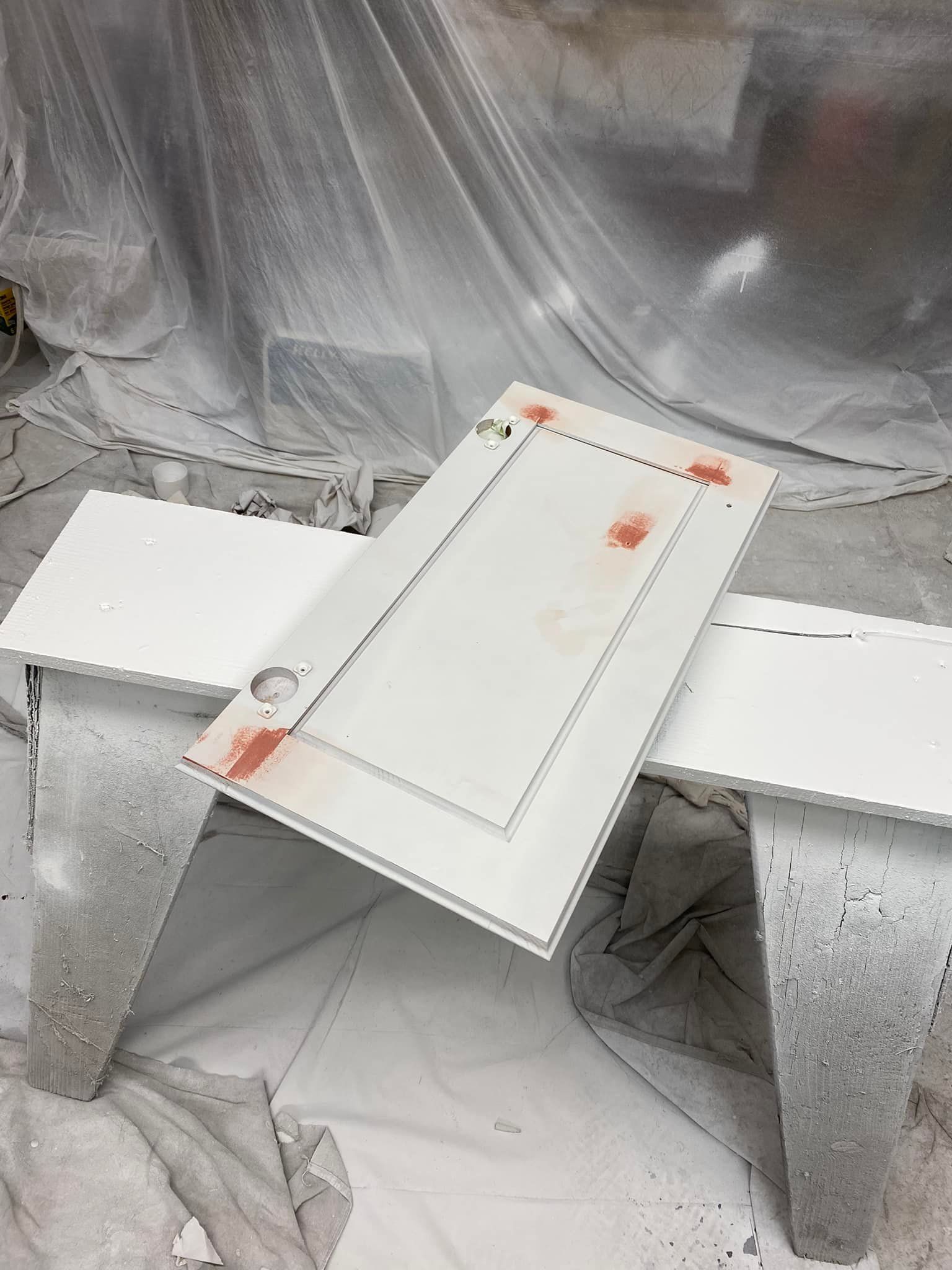 White cabinet door with pinkish spots, propped on a white painted sawhorse in a paint booth.