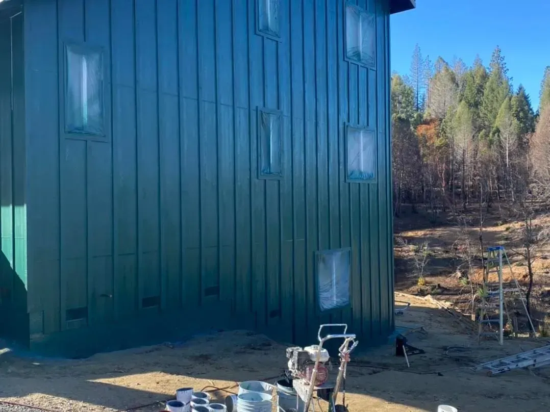 Green house exterior being painted, windows covered. Paint sprayer and supplies on the ground. Forest backdrop.
