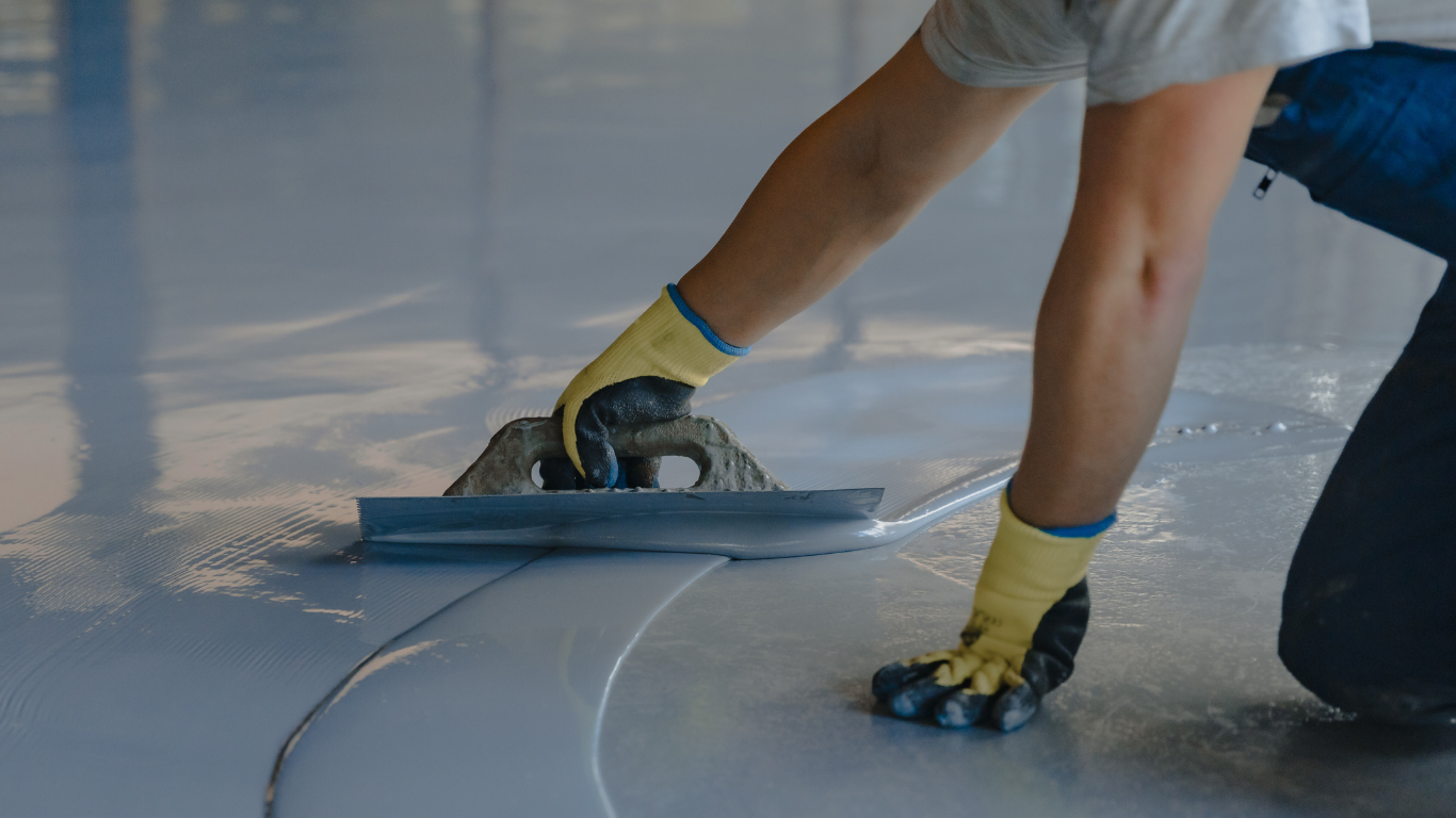 Person kneeling, smoothing light gray epoxy flooring with a trowel, wearing gloves.