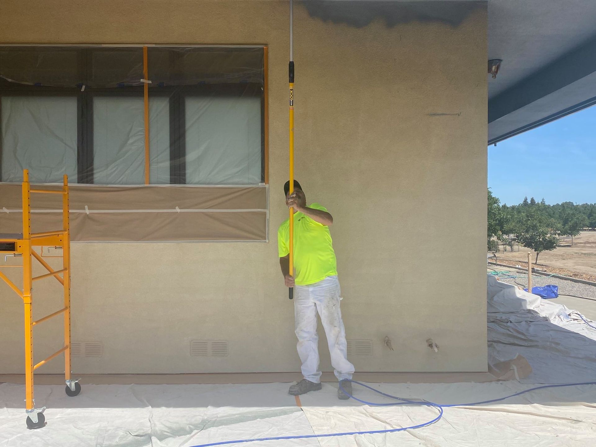 A person painting exterior wall with an extension pole; standing on a covered patio near a window.