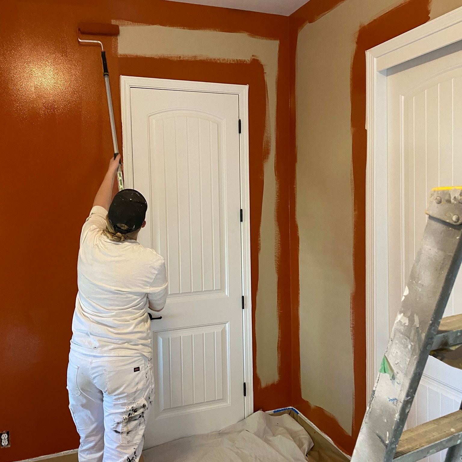 Person painting an interior wall terracotta, using a roller. Door and adjacent walls are beige.
