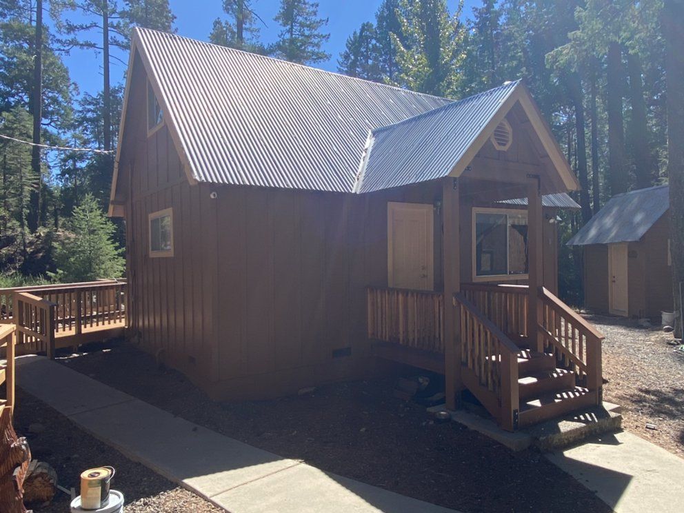 Brown cabin with a metal roof and wooden porch, set in a wooded area.