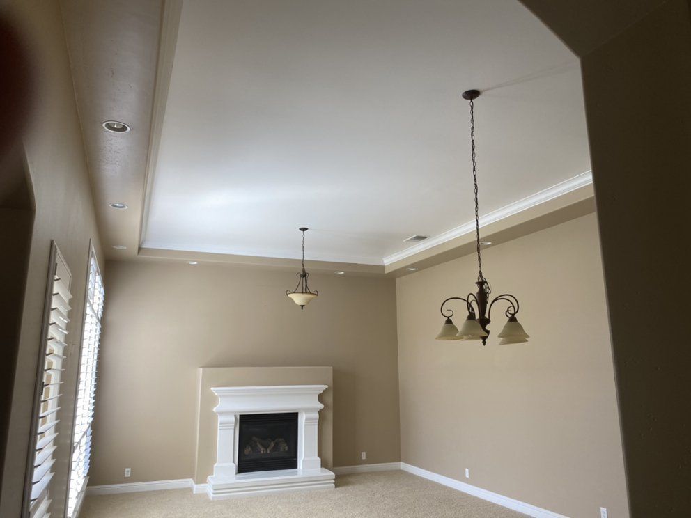 Interior view of a living room with a fireplace, two chandeliers, and large windows with shutters; walls are beige.