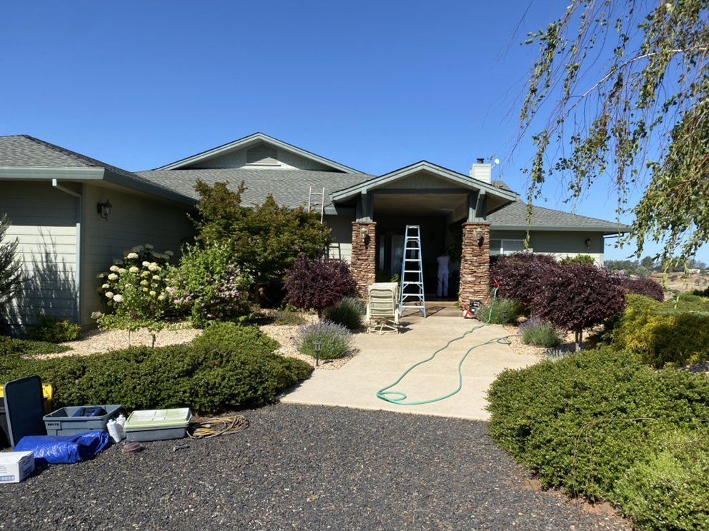 A house exterior with landscaping and a ladder by the entrance. Green and tan tones, sunny day.