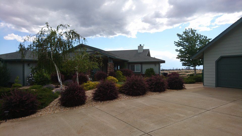 House with gray siding and driveway, landscaping with red bushes and rocks, under a cloudy sky.