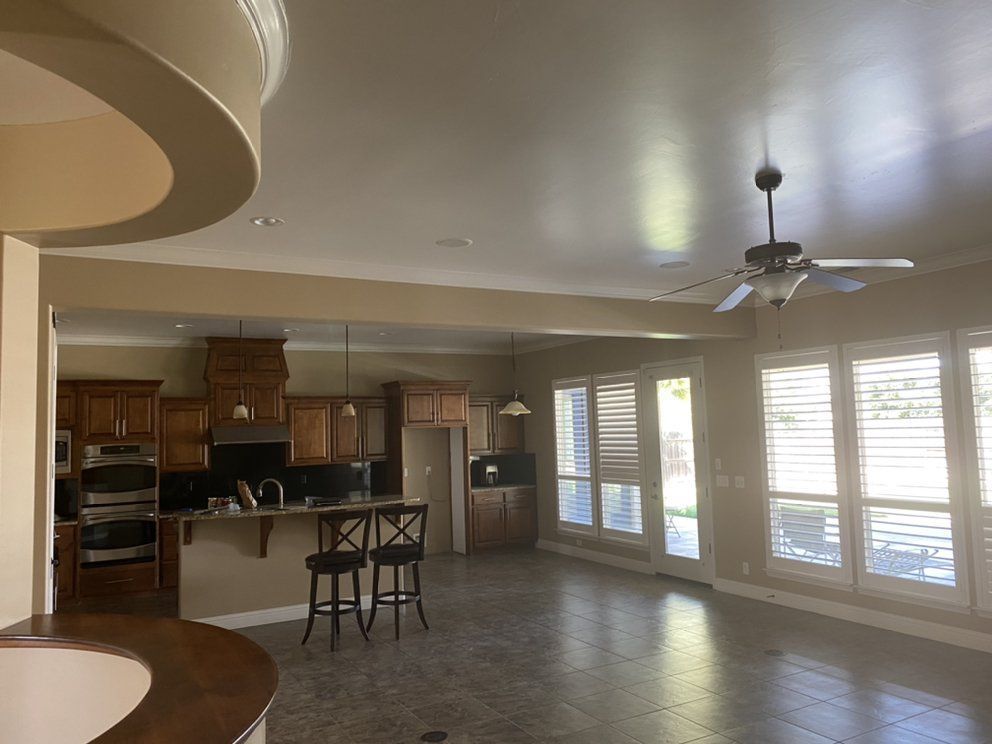 Open kitchen with wooden cabinets, island with stools, and large windows with blinds.