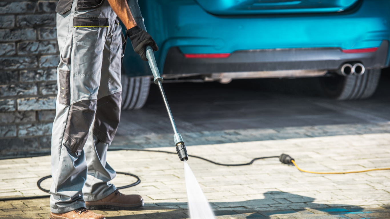 Person wearing work clothes power washing a turquoise car outdoors.