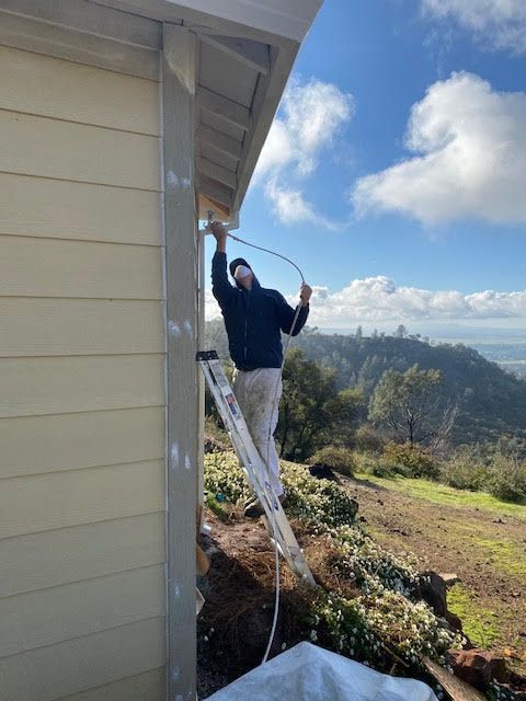 A person on a ladder is working on the side of a building, holding a hose. Sunny, outdoor setting.