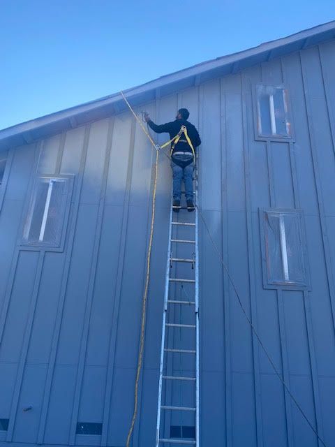 Person on ladder working on the roofline of a blue building, secured by a safety harness.