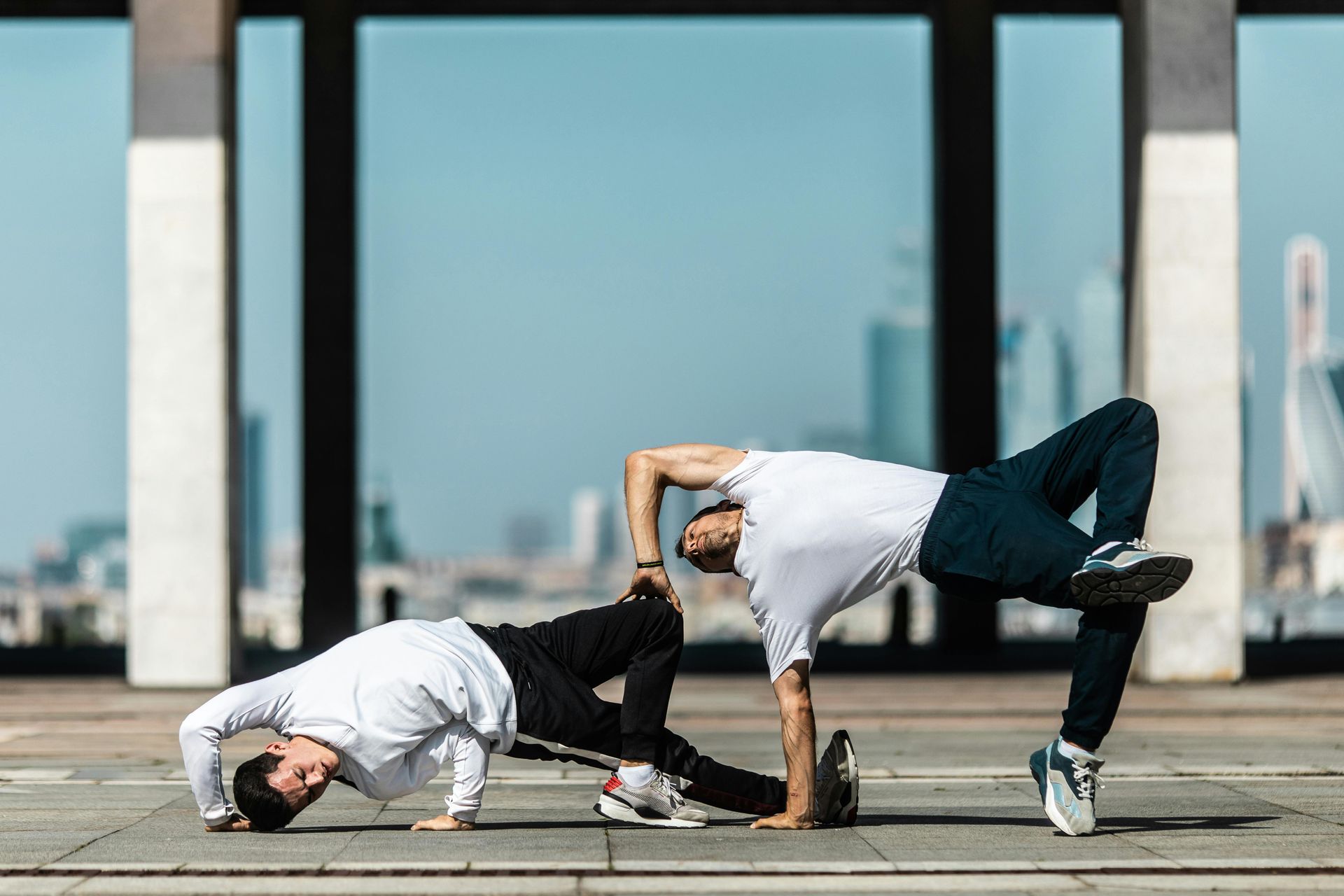 Two dancers performing a breakdancing move on a plaza, city background.