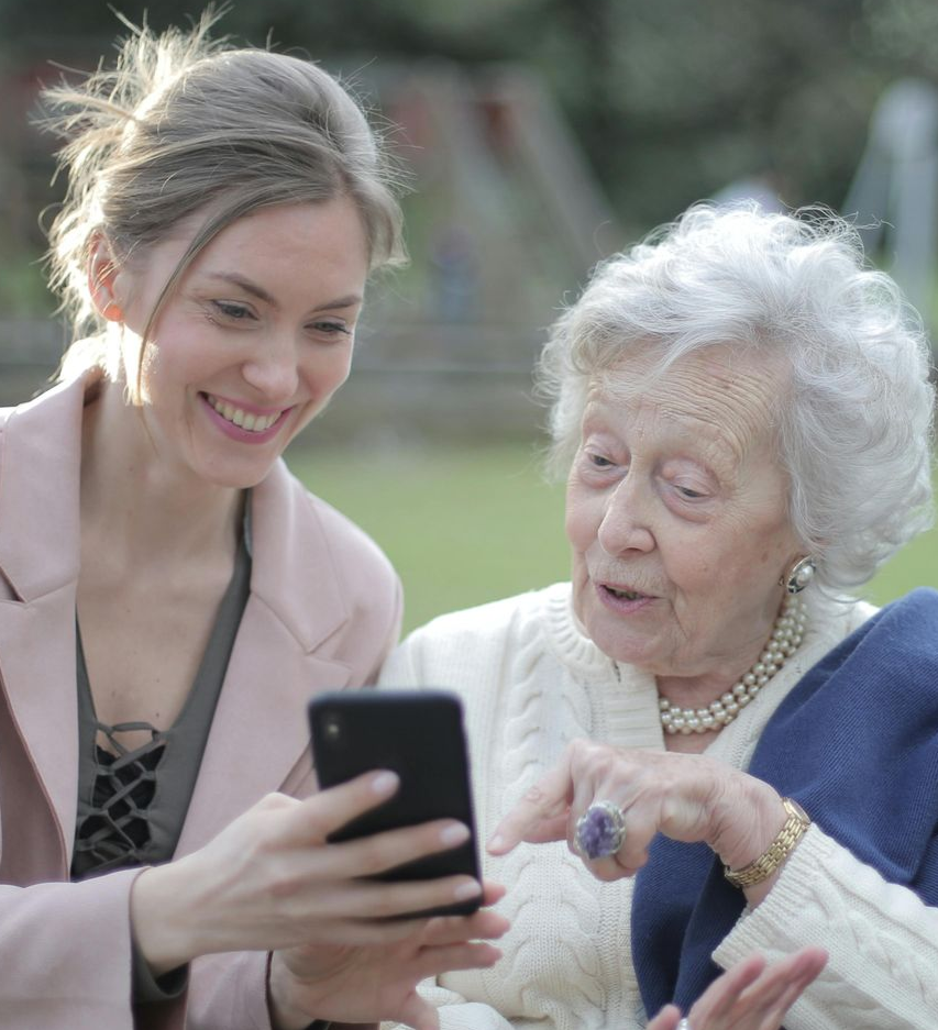 Young woman shows phone to elderly woman outdoors; both smile and look at the screen.
