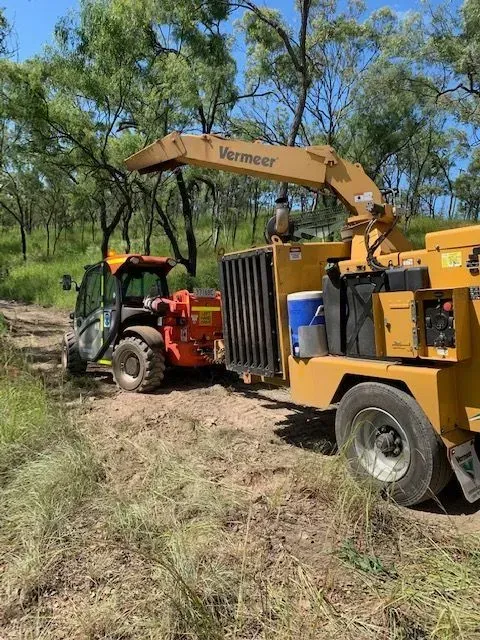 Yellow wood chipper towed by an orange tractor in a wooded area — Boyne Island Tree Services in Boyne Island, QLD